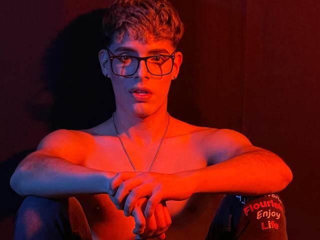 AngelEvil with curly blonde hair, glasses, and red lighting, shirtless, arms crossed, looking at camera, wearing a chain necklace, sitting against dark background.