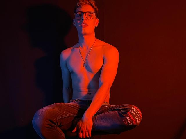 Shirtless AngelEvil with glasses, red light, and a dog tag necklace, sitting cross-legged in dark jeans, showing a toned, muscular, and cute upper body.