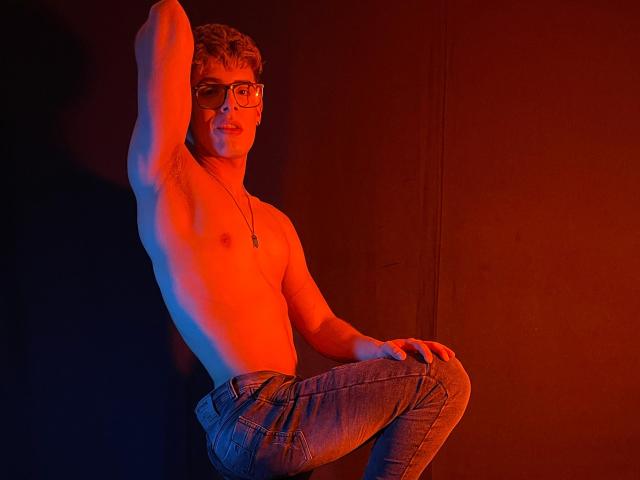 Shirtless AngelEvil with glasses, blue eyes, and short brown hair, sits against a dark wall, wearing tight jeans, red and blue lights highlighting their muscular, bare chest.