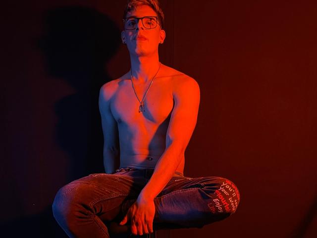 Shirtless AngelEvil with glasses, red light, sitting cross-legged, wearing jeans, necklace, and slight shadow on dark background. Hands on knees, relaxed, with visible chest and jeans.