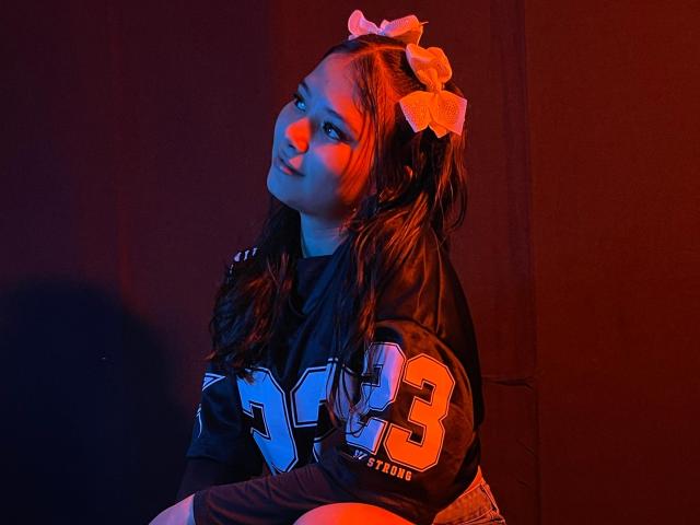 LizzieSweets with wavy brown hair, wearing a black NFL jersey with orange numbers, white bows in hair, sitting, looking up with a slight smile, red and blue lighting.