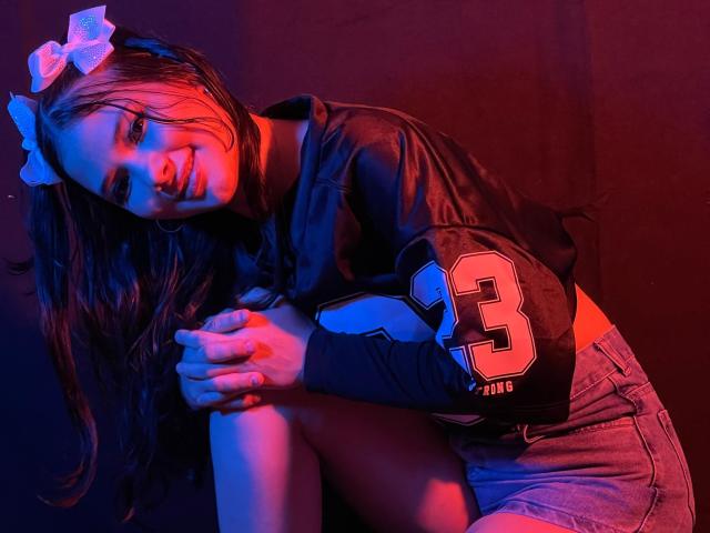LizzieSweets with long brown hair, white bow, black sports shirt, number 3, denim shorts, sitting, smiling, hands on knee, red and blue lighting, looking at camera.