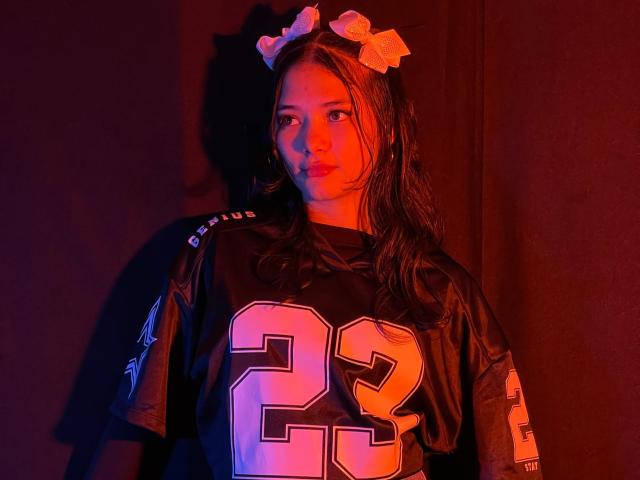 LizzieSweets wearing a black NFL jersey with number 23, has wet brown hair, pink bow headband, and red-orange lighting, standing against a dark background.