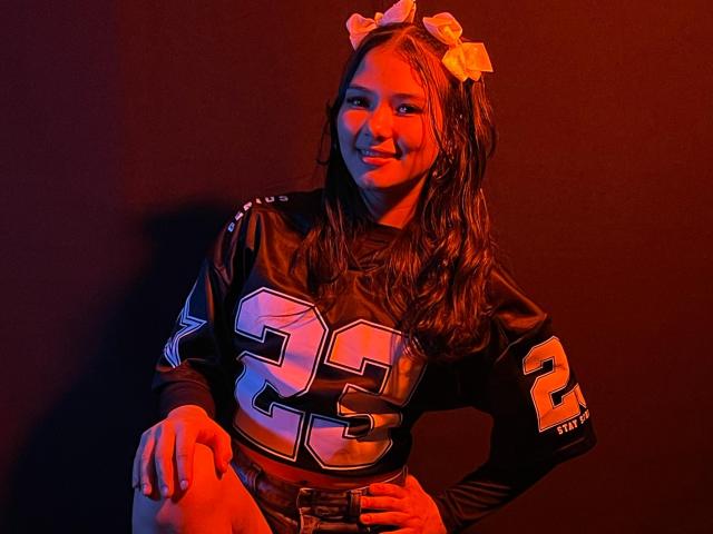 LizzieSweets, with wet brown hair, wears a black football jersey numbered 25, and a yellow bow, sitting, smiling, red and orange lighting, hands on knees, looking cute.