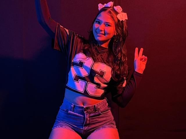 LizzieSweets with long brown hair, wearing a black football jersey number 25, denim shorts, and a white bow, making a peace sign, and smiling in red and blue lighting.
