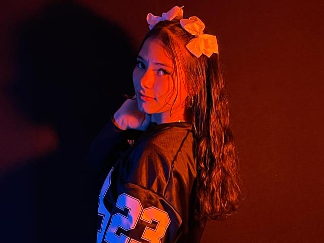 LizzieSweets with long brown hair in pigtails, wearing orange bow, black football jersey, looking over shoulder, holding chin, shadowed in red-orange light, showing cute, sexy face.