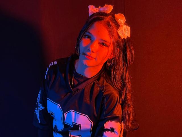 LizzieSweets with red-orange lighting, wearing a black NFL football jersey, number 97, and orange hair bows, has long brown hair in a ponytail, smiling subtly.