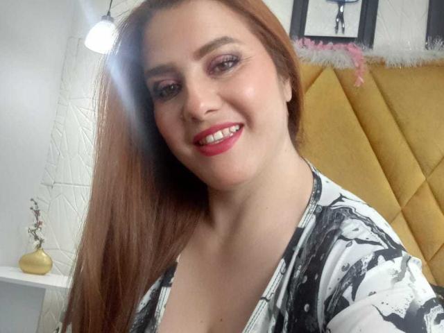 Freya mit langen, braunen Haaren, rosa Lidschatten, rotem Lippenstift, lächelnd, weißes und schwarzes Blumenhemd, gelbes Kopfteil, Licht im Hintergrund, helle, weiße Wand, Licht, das auf ihr Gesicht fällt..