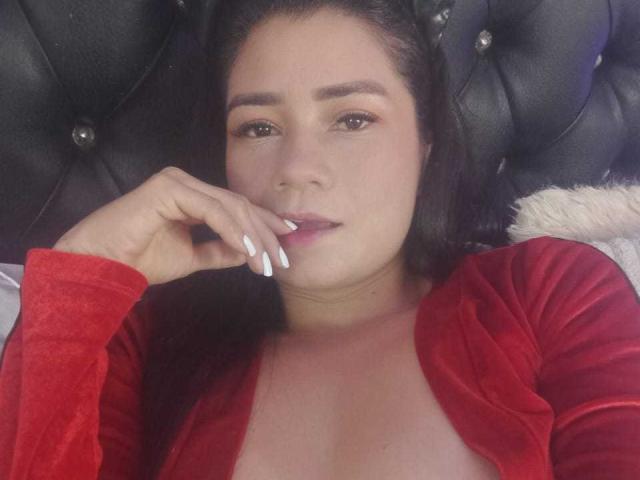 MelissaRonnie with medium brown skin, dark hair, and white nails, wearing a red cardigan, slightly open to show her tits, lying on a black tufted headboard.