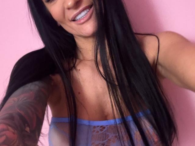 AndraD with long black hair, smiling, showing teeth, wearing a blue lace top, exposing small tits, and a tattoo on her right arm. Pink background.