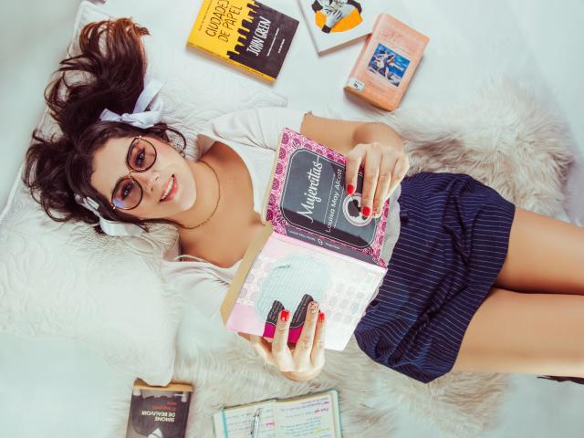GoddessBar lying on a furry white rug, wearing glasses, white top, blue pinstriped skirt, reading a pink-covered book, with colorful books and a tablet around her. Boobs visible.