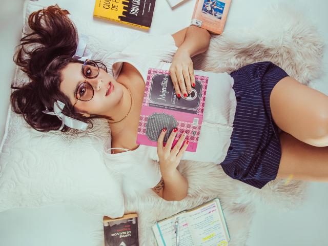 GoddessBar with glasses, lying on a fluffy white rug, wearing a white ribbed top, blue striped skirt, reading a pink book, surrounded by books, with red nails.