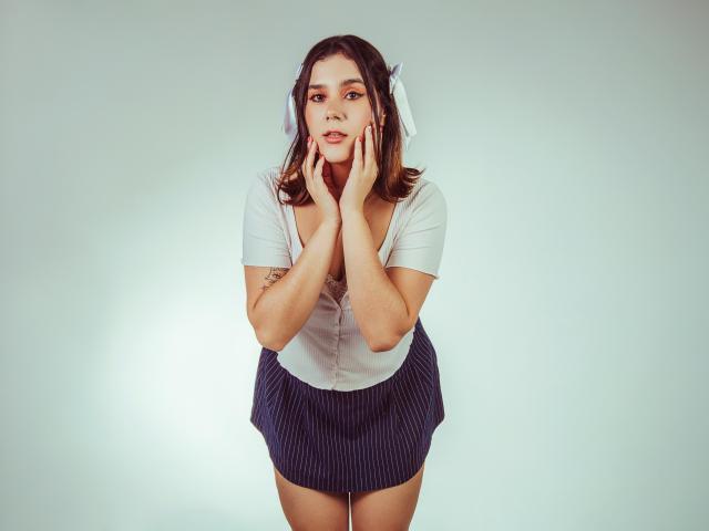 GoddessBar has medium tits, brown hair, and a white shirt with a pinstripe skirt, hands on her face, looking cute with a blue hairpin, standing against a plain background.