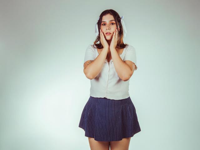 GoddessBar with hands on cheeks, white short-sleeve shirt, blue pinstripe skirt, brown hair, looking surprised, standing against a plain background. Boobs, ass, pussy not visible.