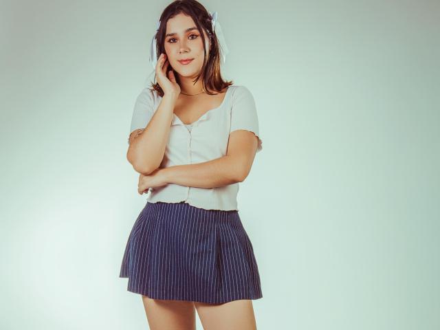 GoddessBar with brown hair, white short-sleeve shirt, and blue pinstriped skirt, touching her cheek, smiling slightly, arms crossed. She's cute with a small ass.