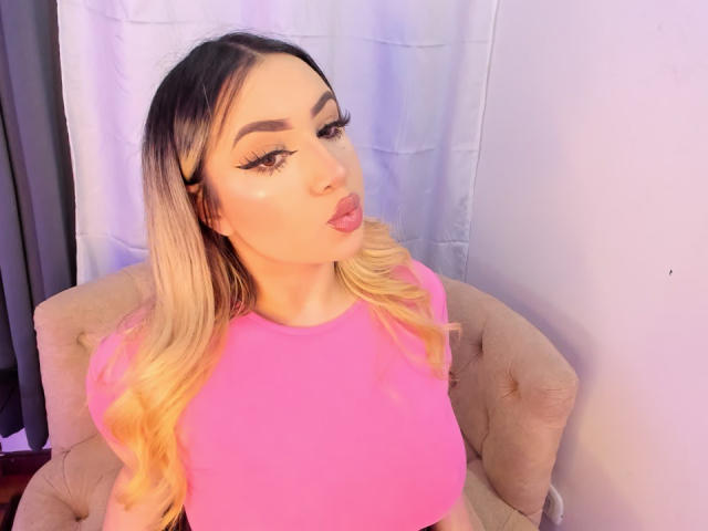 MelissaLust with long blonde hair, pink top, and full lips, pouting slightly, sitting on a beige chair, wearing dramatic eyeliner and mascara, with a white curtain behind her.