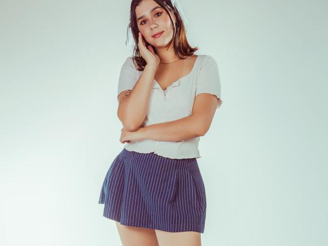 GoddessBar with small tits, wearing a white shirt and blue pinstriped skirt, standing with one hand on her head, smiling, light skin, brown hair, tattoo on her arm.
