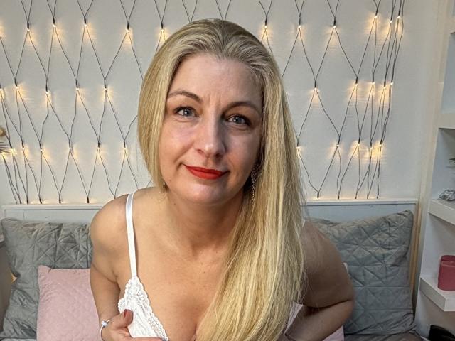 Blonde MissyNia with red lipstick, wearing a white lace top, smiling at the camera, holding her tits gently, sitting on a gray quilted bed, with fairy lights in background.