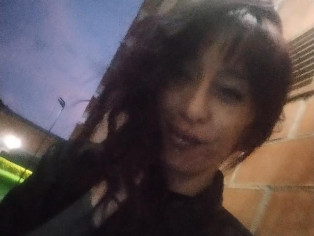 A blurry photograph of GrethelMulher with dark, wavy hair and light skin. She's slightly smiling with her lips parted. Her face is partially obscured by her hair. She's wearing a black shirt. The background has a brick wall on the right and a green light on the left. The image has a low-light, grainy texture, with some blue light visible in the upper left. The composition is slightly off-center, focusing on GrethelMulher's face.