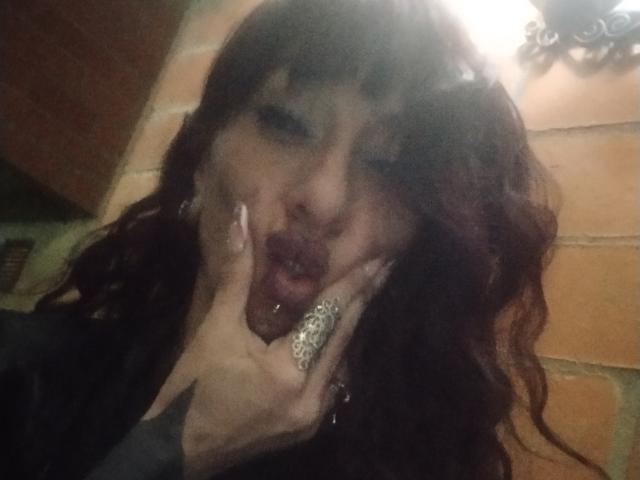 GrethelMulher has dark, wavy hair and is making a dramatic, open-mouthed facial expression. She holds her face with both hands, her fingers adorned with a large, ornate ring. Her lips are parted, showing a small, metallic lip piercing. She has dark makeup and long, white-painted nails. The image is slightly blurry with a warm, orange-toned background. The composition is close-up, focusing on GrethelMulher's face and hands.