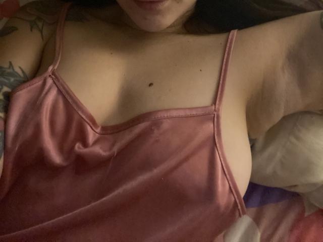 IsabellaPalacio's pink camisole, showing her big tits with a small mole above, and a tattoo on her left arm. Her arm is raised, and she's lying down.