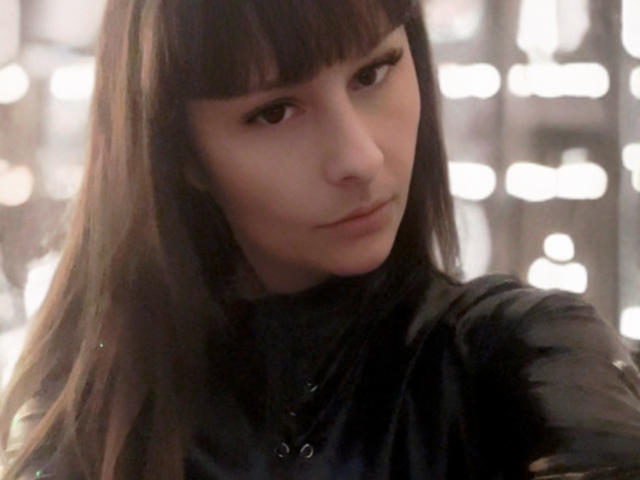 DeesseHera with dark brown hair, straight bangs, and a serious expression, wearing a shiny black leather jacket, looking slightly to the side. Soft light highlights her face.