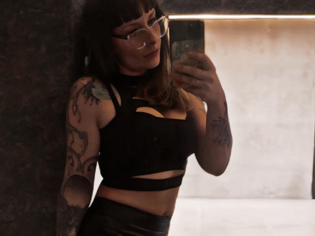 DeesseHera with glasses, black cutout top, showing her tits, tattoos on arms, taking a mirror selfie, wearing black leather skirt, holding phone, dark hair, confident smile.