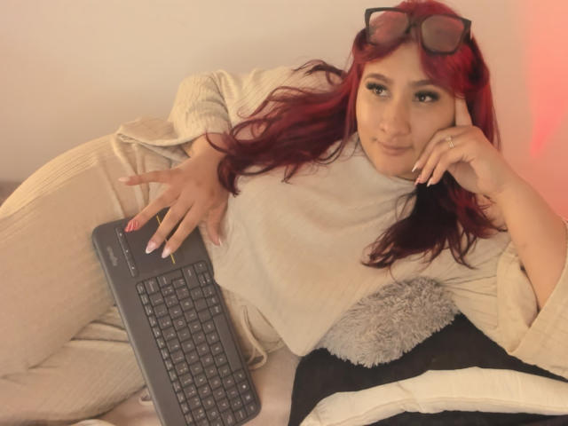 ElyRoss with red hair, wearing glasses on head, beige sweater, and pants, lying on bed, using black laptop, smiling, fingers with pink nail polish, soft light, relaxed.