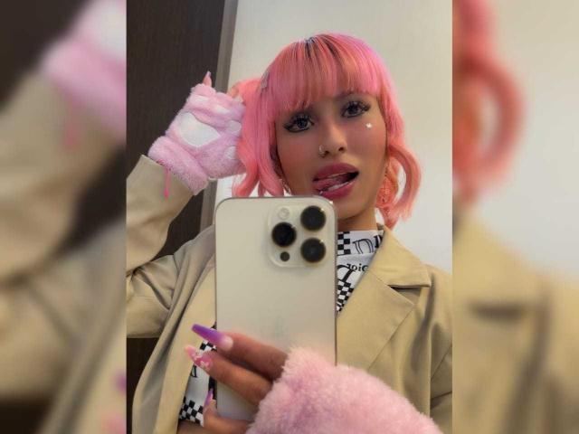 MilaRouge with pink hair, tongue out, wearing pink gloves and beige coat, holding iPhone, taking selfie in mirror, showing her ass in reflection, purple nails, cute expression.