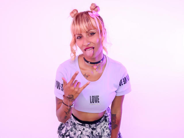 MilaRouge with blonde double buns, pink bow, tongue out, white crop top, "LOVE" text, tattoos, choker, "WELCOME" text, making a rock-and-roll hand sign, purple pink lighting.