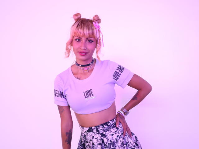 MilaRouge has pink-dyed hair in buns, wearing a white crop top, black choker, and floral skirt, with tattoos on her arms and a cute pink hairclip.
