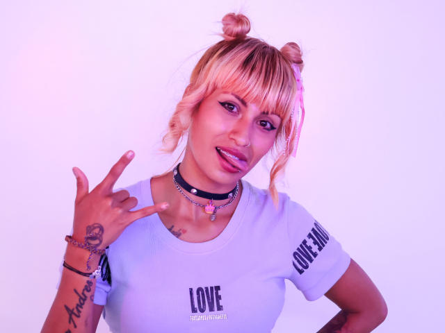 MilaRouge with pink-tinted blonde hair in buns, wearing a white shirt, black choker, and pink lipstick, sticking out tongue, making a peace sign with tattooed hand.