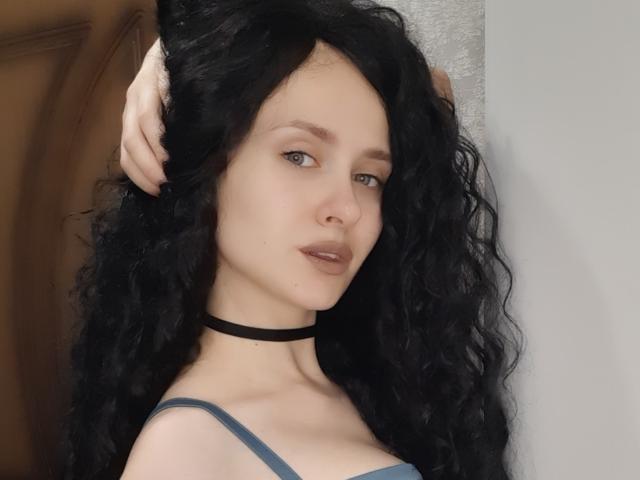 VictoriaAlarie with black curly hair, pale skin, blue eyes, wearing a black choker and blue strap top, touching her head, showing a subtle, sexy expression.