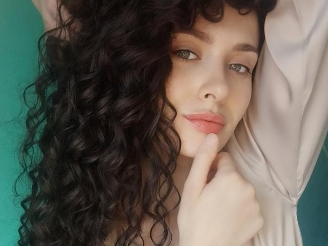 VictoriaAlarie with long curly black hair, pale skin, green eyes, pink glossy lips, and a finger touching her mouth, wearing a beige shirt, looking slightly to the side.