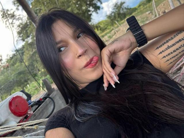CloeThonsom has long black hair, glossy pink lips, and a tattoo on her right arm. She's wearing a black shirt, looking to the side, with a black wristwatch.
