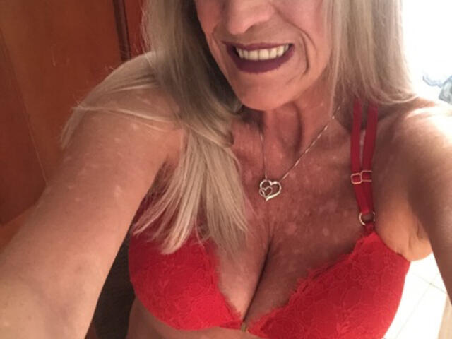 Jen, met lang blond haar, lachend, draagt een rode kantbeha die haar grote borsten laat zien, en een hartvormige ketting. Ze heeft donkerpaars lippenstift op en maakt een selfie..