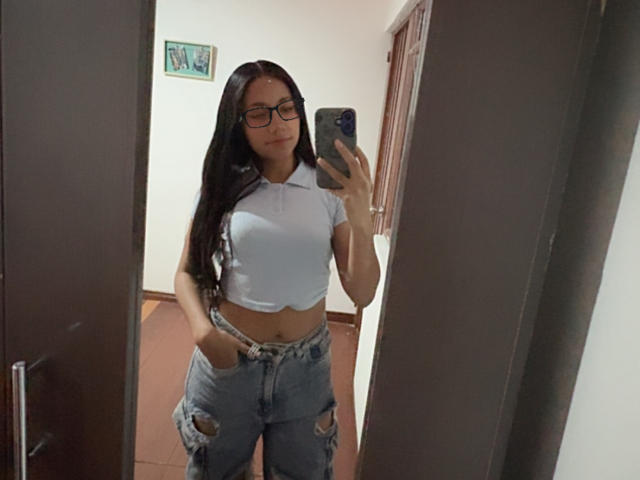 AnaRome with long black hair, glasses, white crop top, and ripped blue jeans, taking a mirror selfie, showing her small tits and hands in her jeans' pockets.