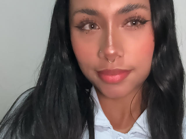AnaRome has long black hair, brown eyes, a nose ring, and pink lips, wearing a white shirt, with subtle freckles and a slight smile.