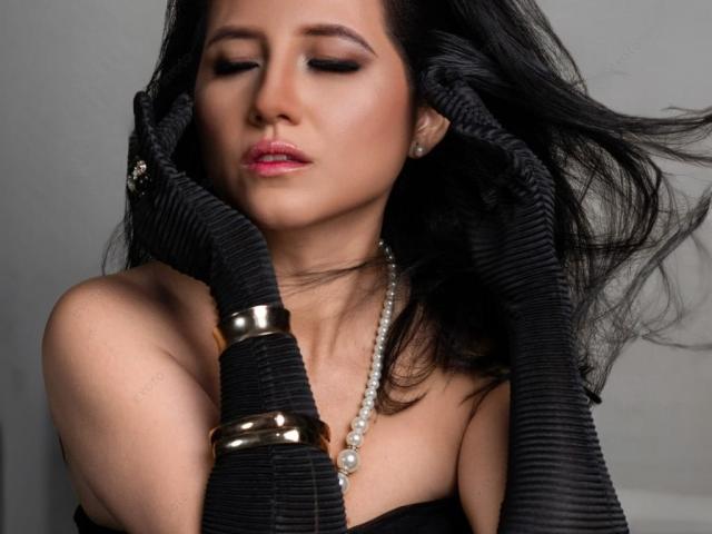 Renata Rivas, met zwarte ribgebreide handschoenen, gouden armbanden, een parelketting en rode lippenstift, die haar hoofd aanraakt met gesloten ogen, waarbij ze haar borsten en haar sexy gezicht laat zien..
