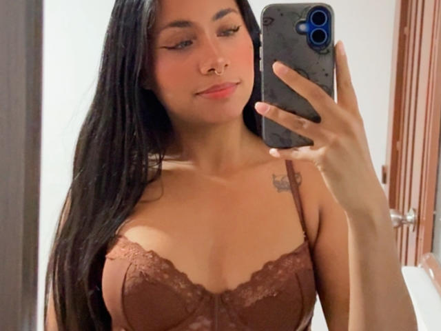 AnaRome with long black hair, wearing a brown lace bra, holding a phone, smiling, showing her tits, nose ring, and tattoo, in a mirror selfie.