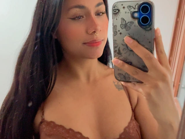 AnaRome, com cabelos longos e escuros, vestindo um sutiã de renda marrom, segurando um celular com capa de borboleta, com um piercing no nariz, maquiagem discreta e uma pequena tatuagem no ombro direito..