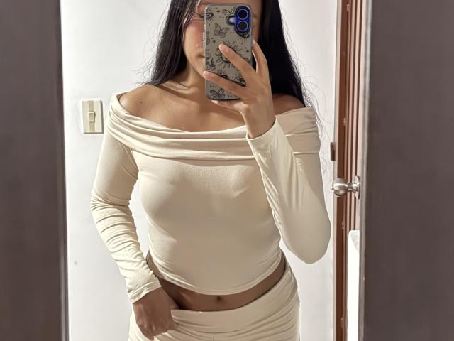 AnaRome in a cream off-shoulder top, showing small tits, with a hand pulling down her cream skirt, holding a phone in a mirror selfie.