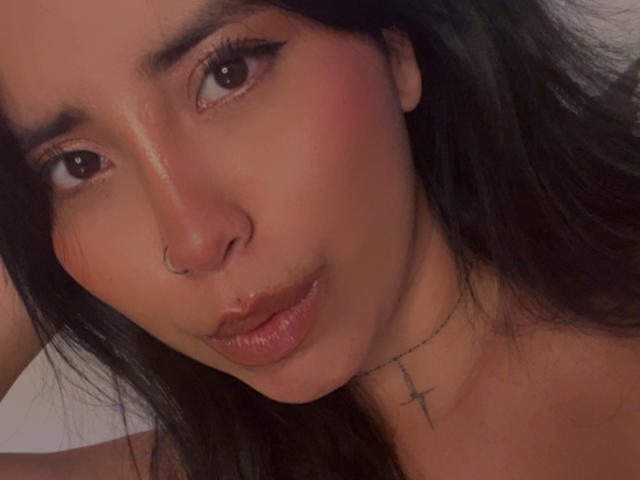 ElizabethMacy's face, nose ring, black hair, brown eyes, subtle blush, small cross necklace, glossy pink lips, slightly parted, looking to the side, soft lighting.