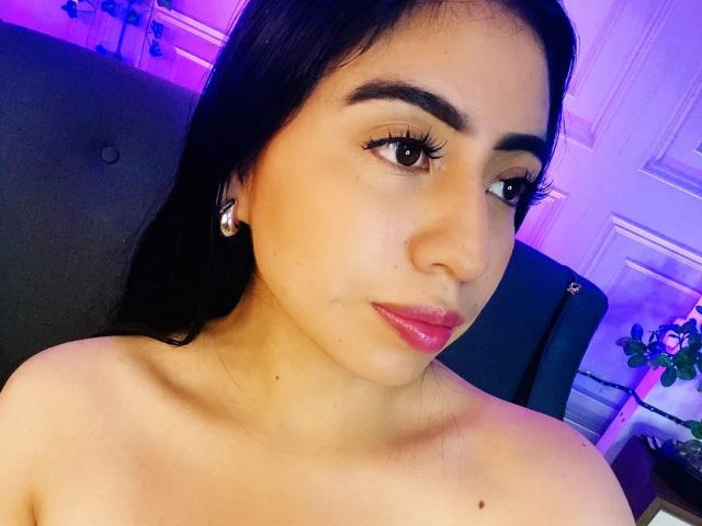 ZoeRedds has tan skin, long black hair, brown eyes, and pink lipstick. She's wearing diamond earrings and looking at the camera with a slight smile, showing her bare shoulders.
