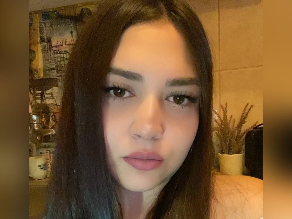 Issabellx has long dark brown hair, thick eyebrows, and pink lipstick, with a neutral expression, looking directly at the camera, in a slightly blurred, warm-toned photo.