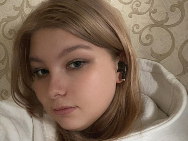 Fastik has light brown hair, blue eyes, and a slight pout. She's wearing black earbuds and a white quilted hoodie, with a subtle, natural makeup look.