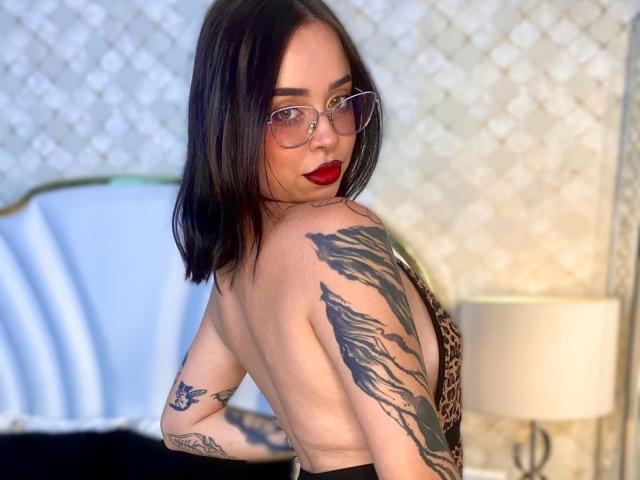 NaiaWhisper with red lips, glasses, black shoulder-length hair, leopard dress, tattoos on arm and shoulder, looking over shoulder, back to camera, showing her ass.