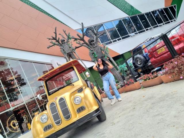 SofiiaThooner stands next to a yellow toy jeep and a red truck, wearing a black top and blue jeans, with long black hair and white sneakers.
