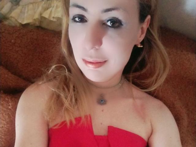 JujuBauva with light skin, blonde hair, and black eyeliner, wearing a red strapless top, small blue gem necklace, slight smile, lying on a brown textured surface.