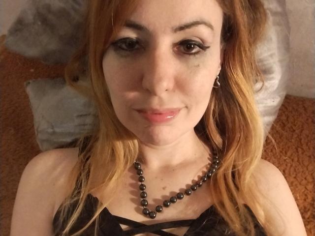 JujuBauva has light skin, long blonde hair, black eyeliner, a black bead necklace, and a subtle smile with a small nose piercing, lying on a pillow.